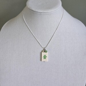 May include: A silver-toned chain necklace featuring a rectangular off-white pendant with a green Chinese character printed on the front. The pendant is accented with a small white pearl and a tiny green bead attached to the silver bail.