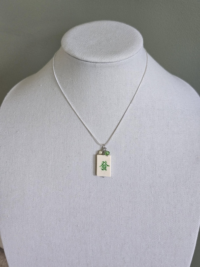 May include: A silver-toned chain necklace featuring a rectangular off-white pendant with a green Chinese character printed on the front. The pendant is accented with a small white pearl and a tiny green bead attached to the silver bail.
