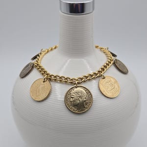 May include: A gold-toned metal chain bracelet featuring multiple dangling coins of various sizes and origins. The coins include a French Republic coin with a profile portrait and a coin marked 20c, all attached to the links of the chain.