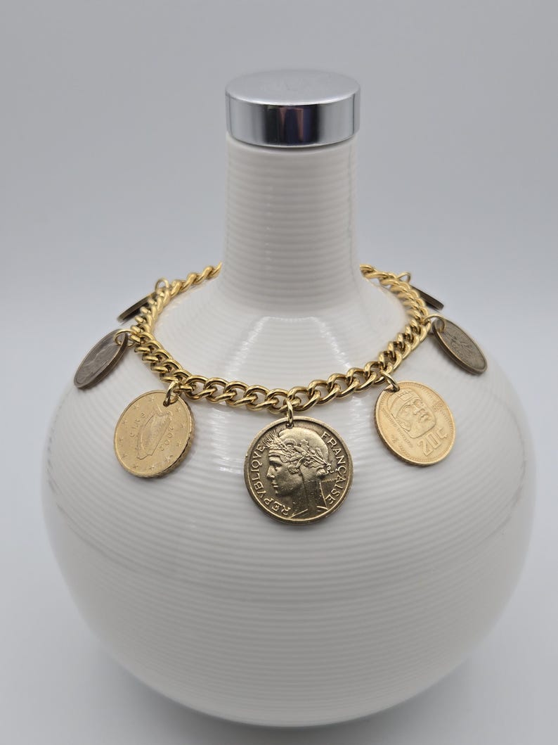 May include: A gold-toned metal chain bracelet featuring multiple dangling coins of various sizes and origins. The coins include a French Republic coin with a profile portrait and a coin marked 20c, all attached to the links of the chain.