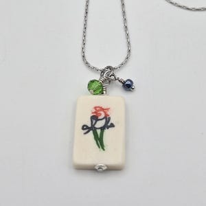 May include: A silver-toned chain necklace featuring a rectangular off-white pendant with a hand-painted bird illustration in red, black, and green. The pendant is accented with a small faceted lime green bead and a tiny dark blue metallic bead.