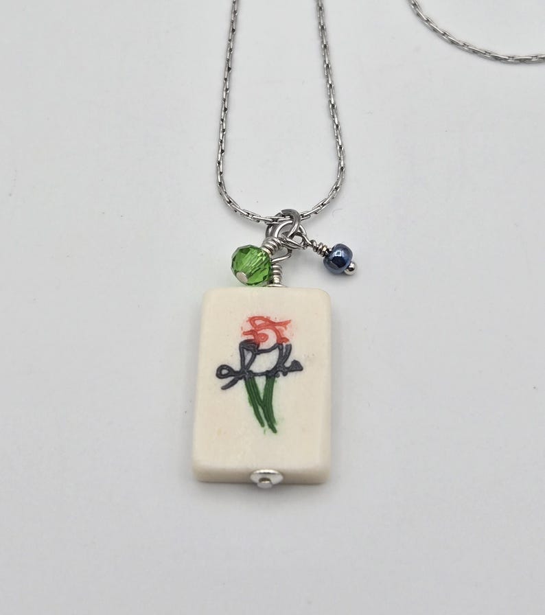 May include: A silver-toned chain necklace featuring a rectangular off-white pendant with a hand-painted bird illustration in red, black, and green. The pendant is accented with a small faceted lime green bead and a tiny dark blue metallic bead.