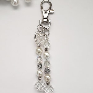 May include: A silver-toned keychain with a lobster clasp, a heart-shaped charm, and dangling strands of white faux pearls, clear faceted beads, and decorative silver-toned beads. The background is white.