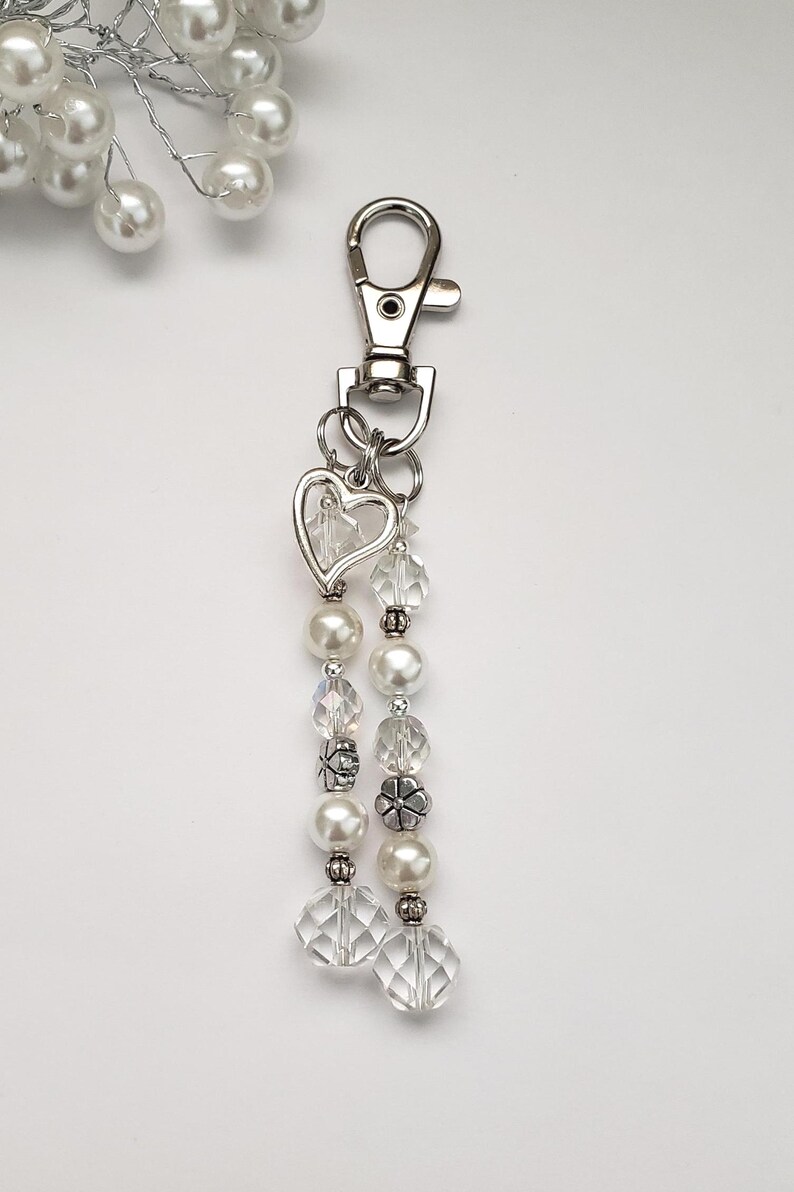 May include: A silver-toned keychain with a lobster clasp, a heart-shaped charm, and dangling strands of white faux pearls, clear faceted beads, and decorative silver-toned beads. The background is white.