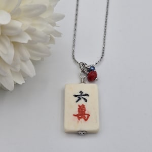 May include: A silver-toned chain necklace featuring a rectangular off-white mahjong tile pendant. The tile displays the black character for six and the red character for ten thousand. A small red faceted bead and a dark blue bead accent the top.