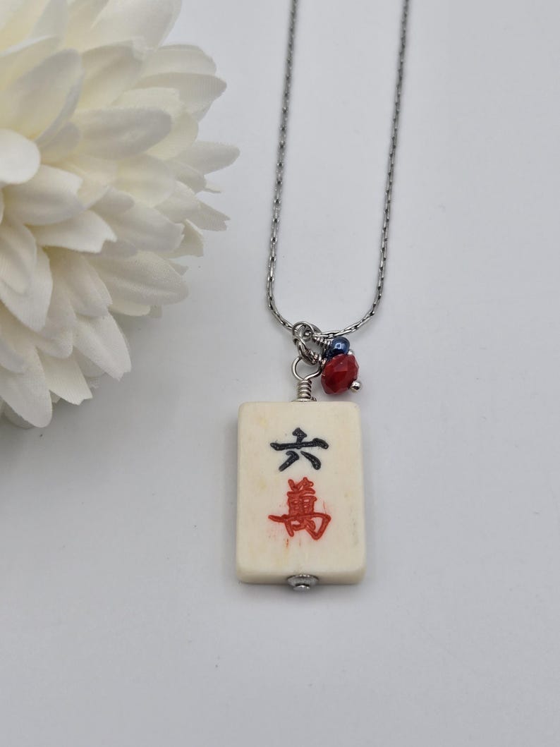 May include: A silver-toned chain necklace featuring a rectangular off-white mahjong tile pendant. The tile displays the black character for six and the red character for ten thousand. A small red faceted bead and a dark blue bead accent the top.
