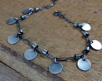 May include: A silver bracelet with a chain and multiple round silver charms. The bracelet is on a wooden surface.