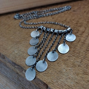 Può includere: Una collana d'argento con una frangia a cascata di dischi rotondi in metallo spazzolato. La collana è realizzata con una catena delicata e ha un design semplice e moderno.