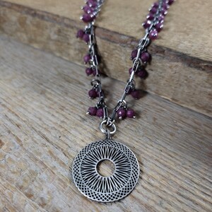 May include: A silver necklace with a large, circular pendant featuring a delicate, intricate design. The necklace is adorned with small, dark pink beads.
