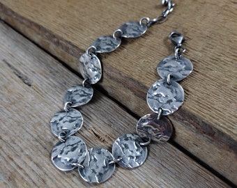 May include: A silver bracelet featuring hammered, circular links. The links are connected by small silver rings and a clasp. The bracelet is displayed on a wooden surface, showcasing its texture and design.