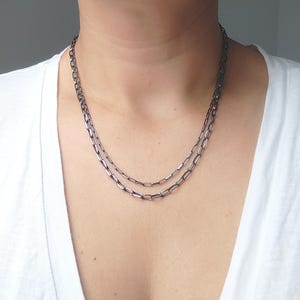 Könnte beinhalten: Eine zweireihige Halskette bestehend aus zwei Strängen mit länglichen, dunkelgrauen, metallisch glänzenden ovalen Gliedern. Die Kette liegt am Hals an und zeigt ein gleichmäßiges Muster aus ineinandergreifenden, rechteckigen Kettengliedern.