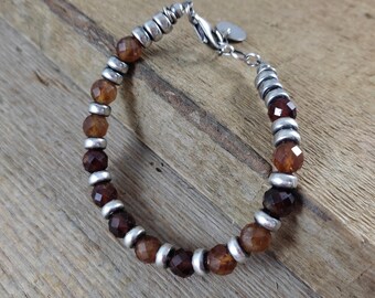 May include: A beaded bracelet featuring alternating sections of faceted, dark brown gemstones and small, silver-colored metal beads. The bracelet has a silver-colored clasp and a small, round charm. The gemstones are a mix of amber and deep brown hues.