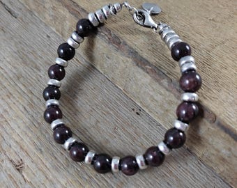 May include: A beaded bracelet featuring dark red, round gemstones and silver-colored metal accents. The bracelet is secured with a silver clasp and a small charm. The bracelet is displayed on a wooden surface.
