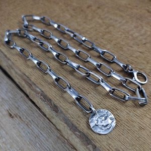 May include: A silver chain necklace with a hammered silver disc pendant. The chain is made of flat, oval links.