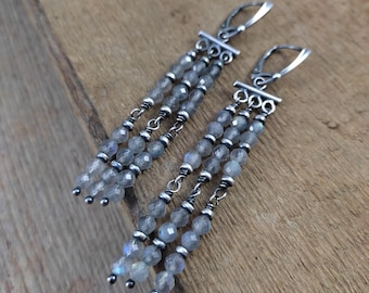 Peut inclure: Une paire de boucles d'oreilles pendantes avec trois rangées de perles grises facettées. Les boucles d'oreilles ont des accents en métal argenté et des fermoirs à levier. Les perles ont une subtile qualité irisée.