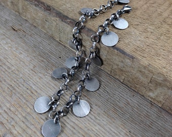 May include: A silver chain bracelet with multiple round silver charms hanging from it. The bracelet is on a wooden surface.