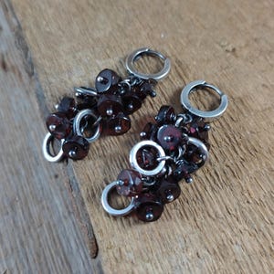 Pode incluir: Um par de brincos de argola de prata com cachos pendurados de contas de pedras preciosas de cor vermelho escuro.