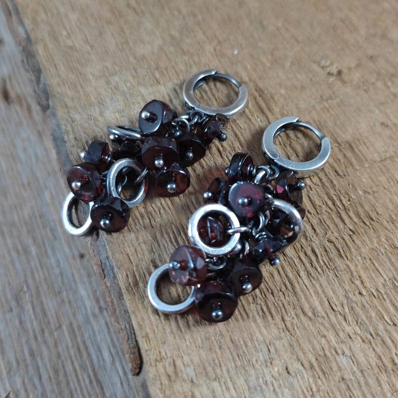 Pode incluir: Um par de brincos de argola de prata com cachos pendurados de contas de pedras preciosas de cor vermelho escuro.