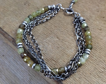May include: A multi-strand bracelet featuring olive-green gemstone beads and multiple silver chains. The bracelet has a lobster clasp closure and is displayed on a wooden surface. The beads are interspersed with silver accents.