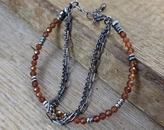 May include: A layered bracelet featuring faceted, reddish-brown beads and a silver chain design. The bracelet has a delicate appearance with multiple strands and silver accents. The beads are small and evenly spaced, creating a refined look.