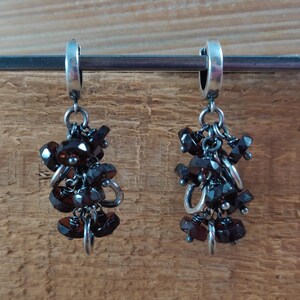 Pode incluir: Um par de brincos de argola de prata com cachos pendentes de pedras preciosas de cor vermelho escuro. Os brincos s&atilde;o feitos de prata e apresentam um design de argola com um cacho pendente de pedras preciosas de cor vermelho escuro.