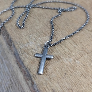 May include: A silver cross pendant necklace. The cross has a simple, classic design with a textured surface, suspended from a matching chain. The necklace is displayed on a wooden surface, contrasting the metal with the wood.
