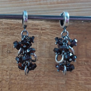 Pode incluir: Um par de brincos pendentes com argolas de prata e um cacho de pedras preciosas castanho-escuras penduradas em cada argola.