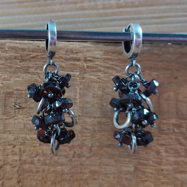 Pode incluir: Um par de brincos pendentes com argolas de prata e um cacho de pedras preciosas castanho-escuras penduradas em cada argola.