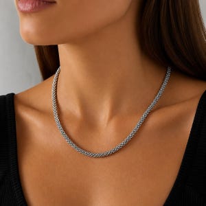 Può includere: Una collana argentata con un design intrecciato. La collana è drappeggiata attorno al collo, mostrando la sua consistenza e lucentezza. Il gioiello è un accessorio classico.