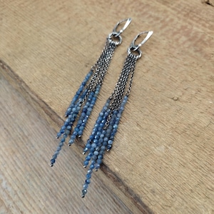 Peut inclure: Paire de boucles d'oreilles pendantes avec des brins de perles bleues suspendus à des chaînes et des crochets d'oreilles en métal argenté. Les boucles sont présentées sur une surface en bois.