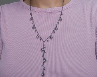 Könnte beinhalten: Eine Y-förmige Halskette aus dunklem Metall mit einer feinen Kette, die mit mehreren glatten, kugelförmigen Perlen unterschiedlicher Größe verziert ist. Die Perlen sind entlang des Ausschnitts angeordnet und bilden einen vertikalen Fall.