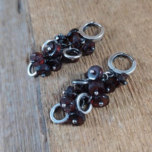 Pode incluir: Um par de brincos de argola de prata com cachos pendurados de contas de pedras preciosas de cor vermelho escuro.