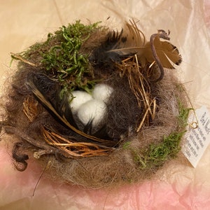 May include: A small bird nest made of twigs, moss, and feathers with three white eggs inside. The nest is sitting on a pink and white tissue paper background. A tag attached to the nest reads "Bird Nests" with text about the nest being made with fiber, twine, and some bits of nature.