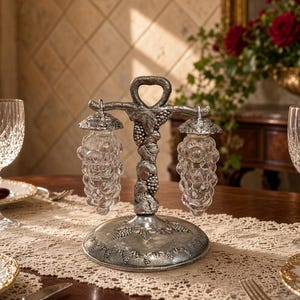 May include: A silver-toned metal stand with a grape vine motif holds two clear glass salt and pepper shakers shaped like clusters of grapes. The stand features a central handle and a round base, both decorated with embossed grape and leaf patterns.