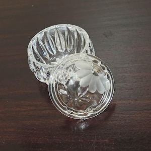 May include: A clear, decorative glass trinket box with a lid. The box has a cut-glass design on the sides and a frosted flower design on the lid. The lid is slightly ajar, revealing the interior of the box. The box is on a dark brown surface.