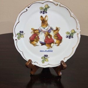 May include: A white porcelain decorative plate with a scalloped edge and gold trim. The center features an illustration of Mrs. Rabbit with three small rabbits in red coats. The text Mrs. Rabbit is printed in blue below the figures, with blackberry clusters.