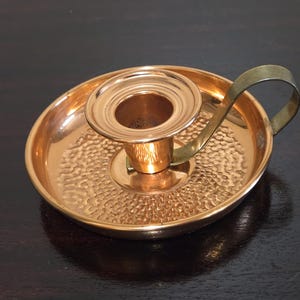May include: A copper-colored candle holder with a hammered texture and a brass handle. The candle holder has a circular base and a central cup to hold a candle. The handle curves upwards from the side.