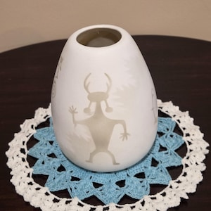 May include: A white, egg-shaped ceramic vase featuring a light brown, stylized graphic of a horned figure with outstretched arms. The vase has a smooth, matte finish and a circular opening at the top, resting on a round, blue and white crocheted doily.