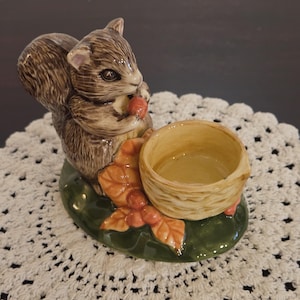 May include: A ceramic squirrel figurine featuring a brown textured coat, holding a small red nut. The squirrel is positioned next to a hollow, light-colored bowl shaped like a woven basket, all set upon a glossy, dark green oval base with orange leaves.