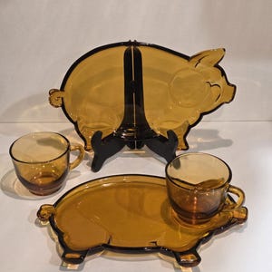 May include: A set of amber-colored glass tableware featuring two pig-shaped plates and two matching glass cups. One plate is displayed upright on a black stand, while the other lies flat with a cup placed on its surface. The glass is translucent.