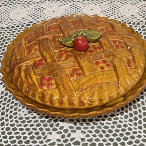May include: Ceramic pie dish with a lattice crust design. The pie dish is glazed in shades of brown and yellow, with red accents. A red apple and green leaves adorn the top. The dish is round and has a scalloped edge.