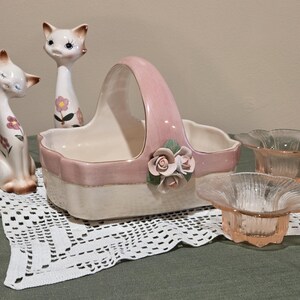 May include: A collection of vintage ceramic decor featuring two cat figurines, a pink and white basket with a handle, and two small pink glass bowls. The cat figurines have floral accents.