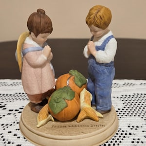May include: A porcelain figurine featuring two children with hands clasped in prayer, standing on a circular base with two orange pumpkins and two ears of corn. The base is inscribed with the text Jessie Willcox Smith in black lettering.