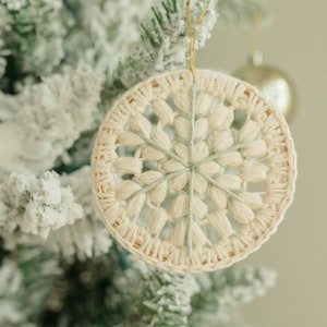 Puede incluir: Un adorno de copo de nieve de crochet blanco y verde colgado de un &aacute;rbol de Navidad nevado.