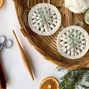 Puede incluir: Dos adornos de &aacute;rbol de Navidad tejidos a crochet en hilo blanco y verde, con un gancho de crochet de madera y un par de tijeras sobre una superficie blanca.