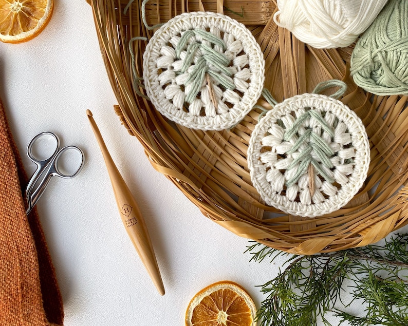 Puede incluir: Dos adornos de &aacute;rbol de Navidad tejidos a crochet en hilo blanco y verde, con un gancho de crochet de madera y un par de tijeras sobre una superficie blanca.