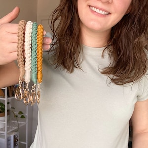 May include: Five colorful braided keychains with gold key rings. The keychains are in shades of brown, cream, green, and yellow.