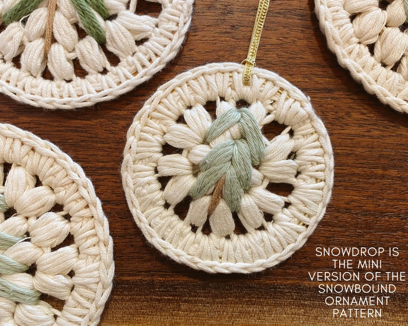 Puede incluir: Primer plano de un adorno de crochet blanco con un dise&ntilde;o bordado en verde y marr&oacute;n. El adorno est&aacute; colgado de una cuerda dorada. El texto "SNOWDROP IS THE MINI VERSION OF THE SNOWBOUND ORNAMENT PATTERN" est&aacute; escrito debajo del adorno.