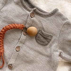 May include: A close-up of a gray baby onesie with a brown wooden teether attached to a burnt orange crocheted strap.