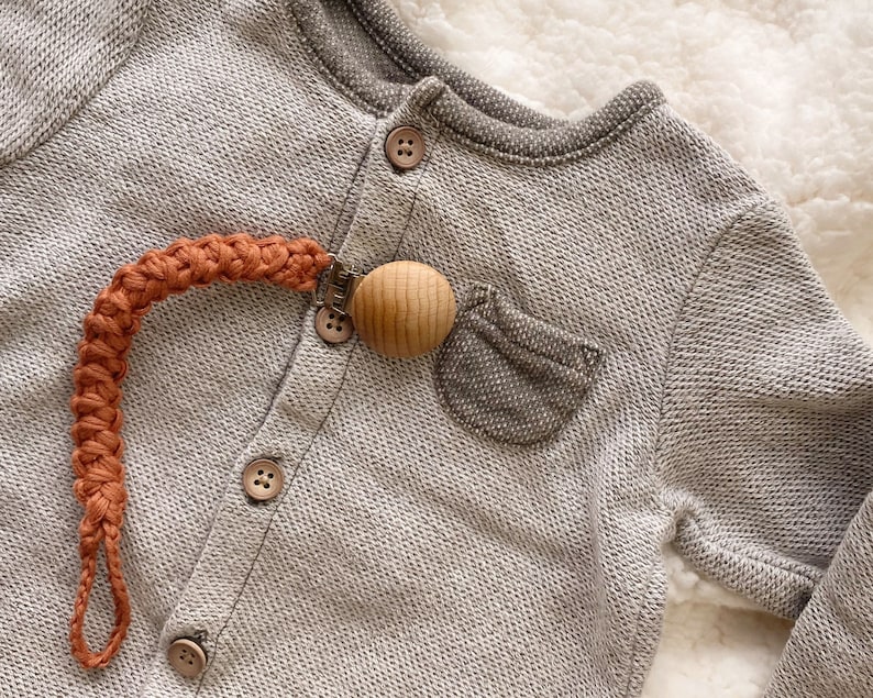 May include: A close-up of a gray baby onesie with a brown wooden teether attached to a burnt orange crocheted strap.